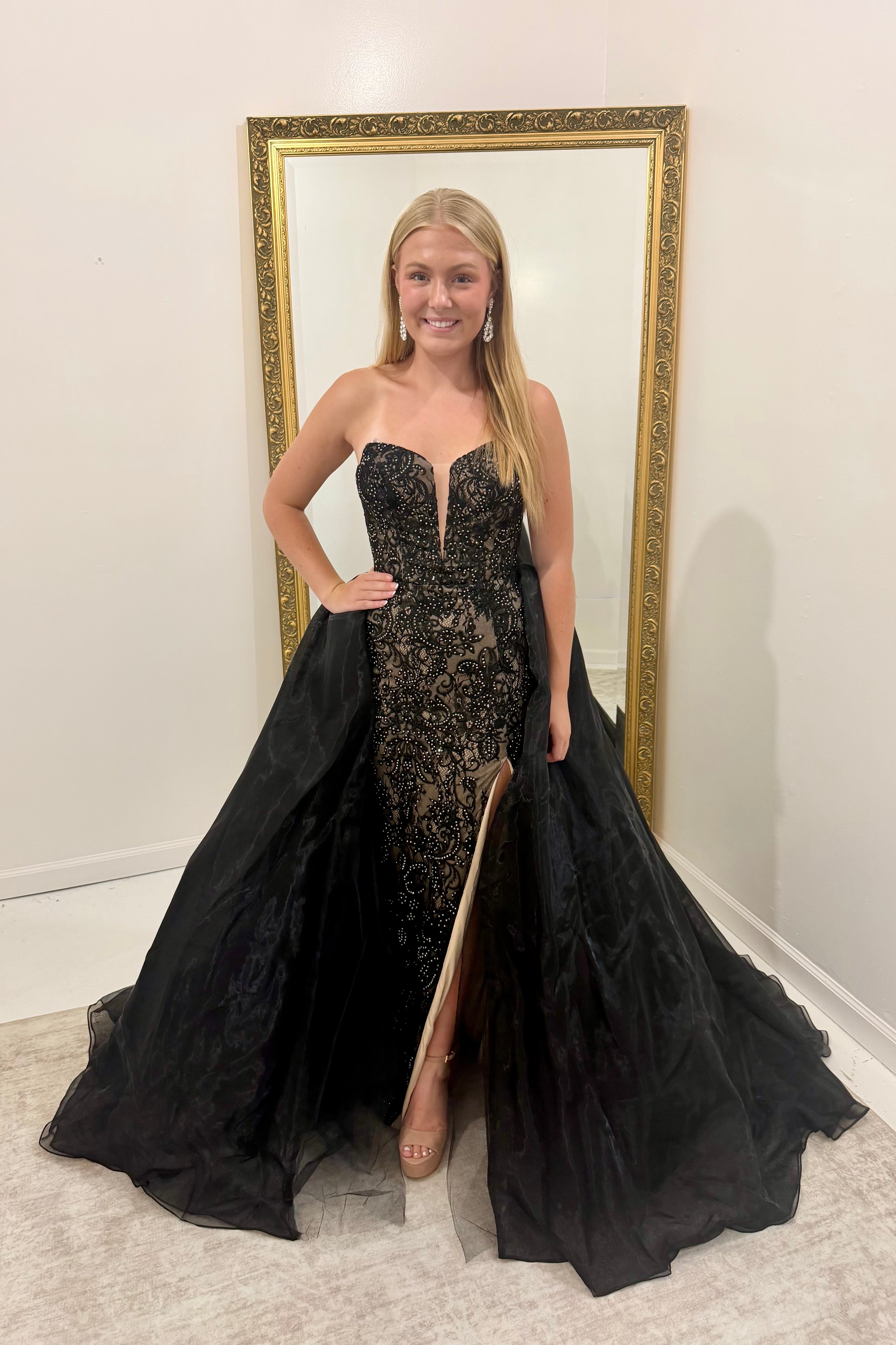 A woman modeling a strapless black lace formal gown with a sweetheart neckline, thigh-high slit, and a dramatic black tulle overskirt, standing in front of a gold-framed mirror.