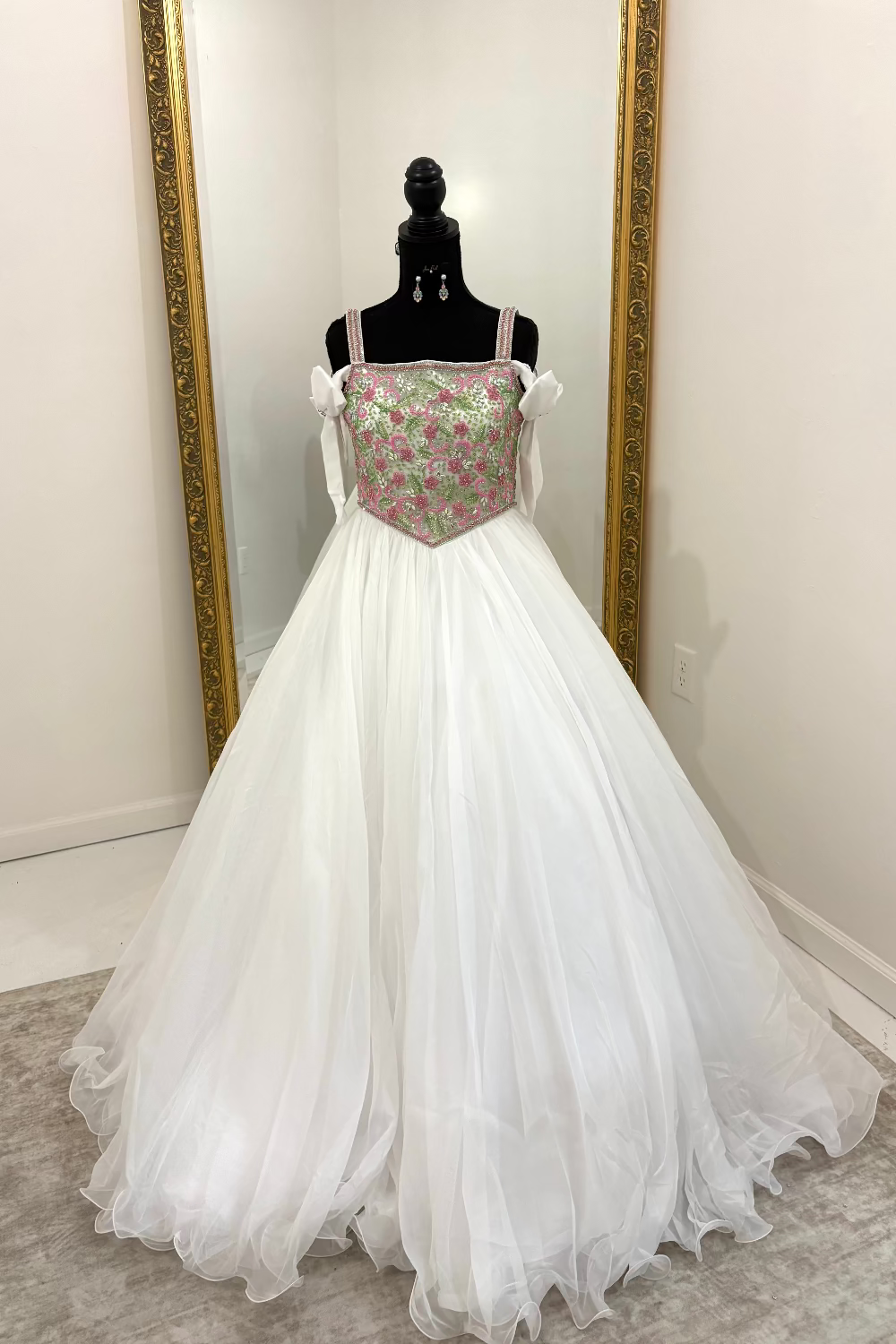 A white ball gown with a pink and green floral embroidered bodice, displayed on a black mannequin in front of a mirror with a gold frame.