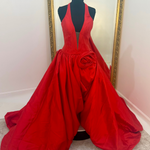 A bright red formal gown displayed on a black mannequin in front of a mirror, featuring a plunging neckline, a large sculptural waist detail, and a voluminous skirt.