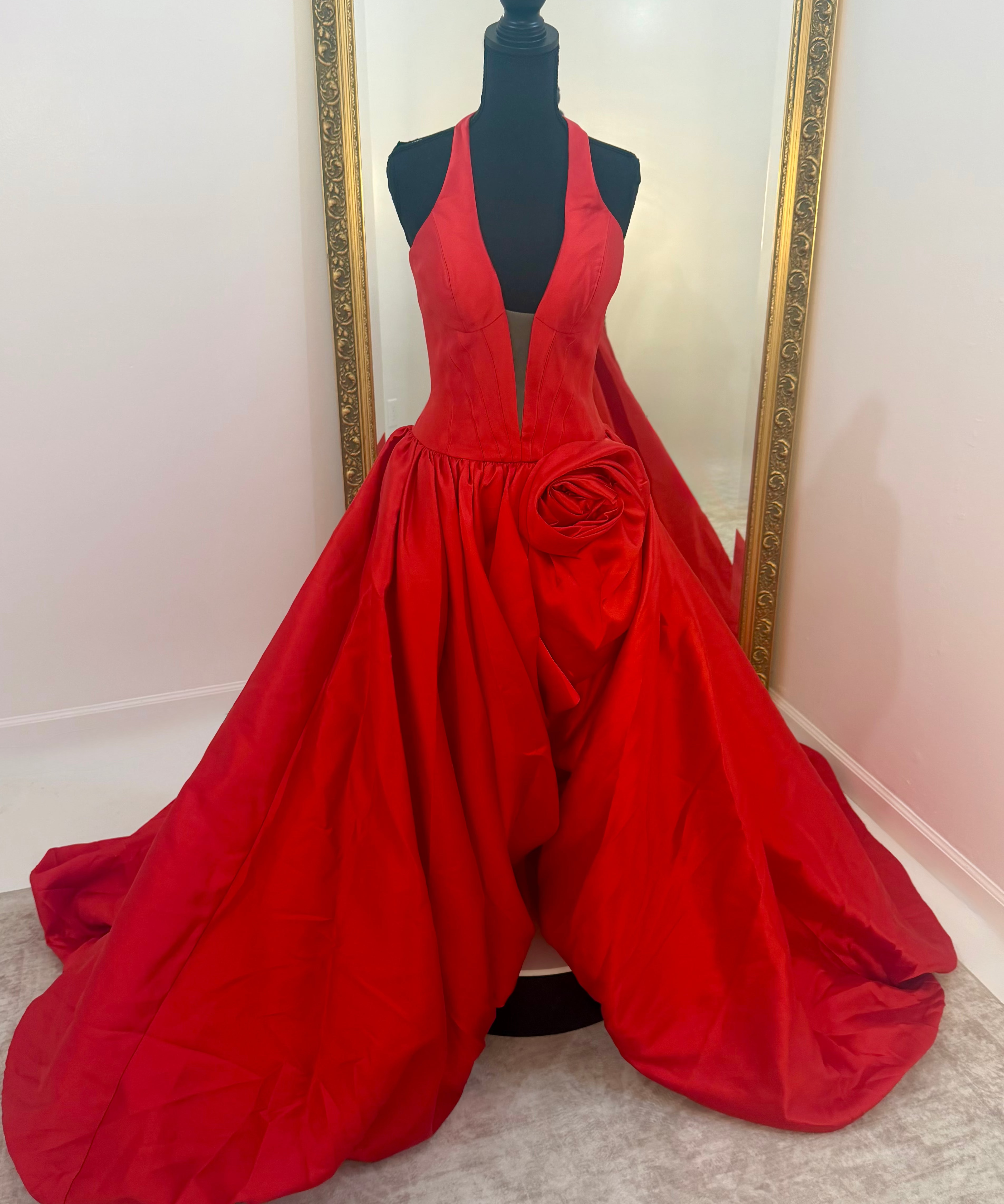A bright red formal gown displayed on a black mannequin in front of a mirror, featuring a plunging neckline, a large sculptural waist detail, and a voluminous skirt.