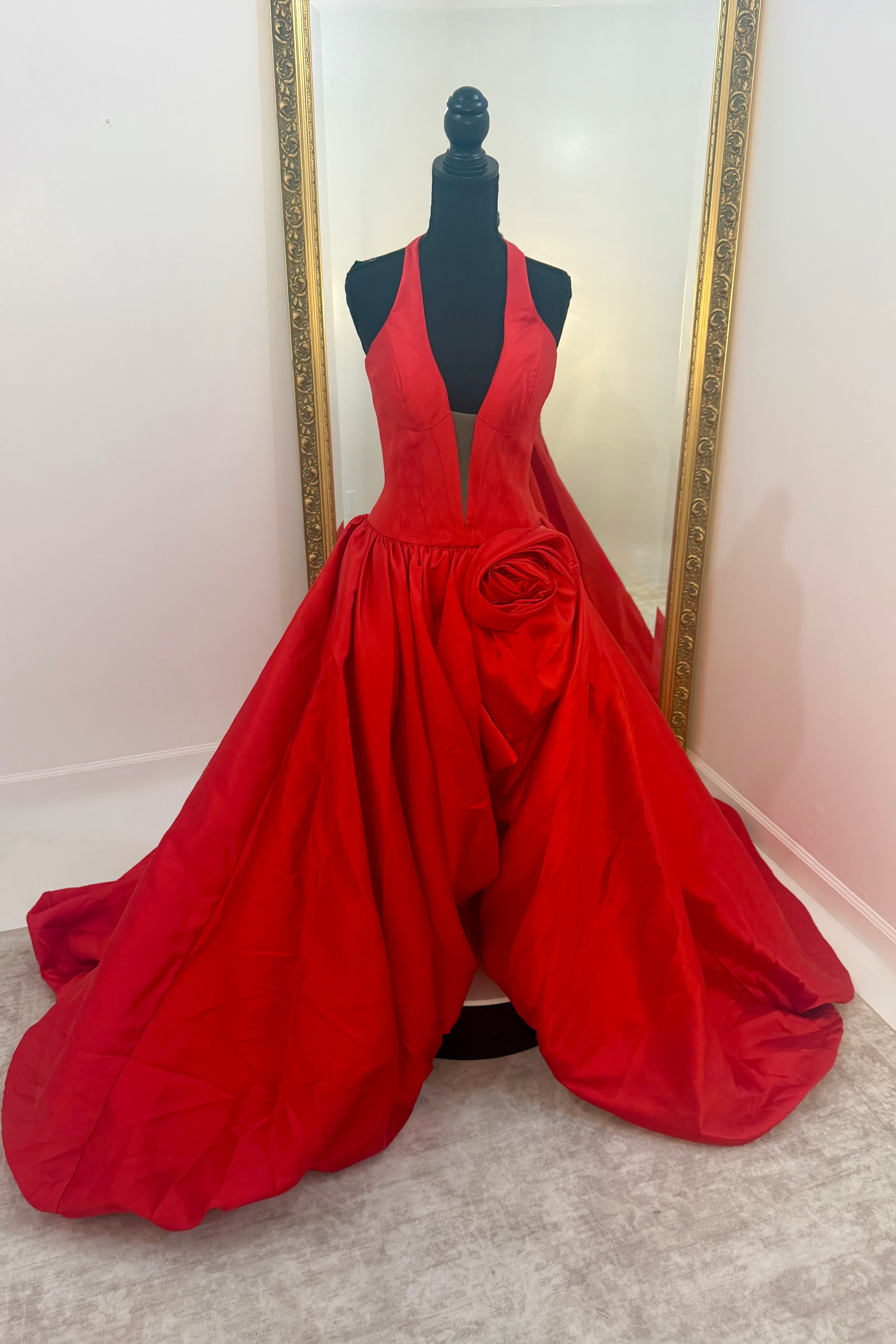 A bright red formal gown displayed on a black mannequin in front of a mirror, featuring a plunging neckline, a large sculptural waist detail, and a voluminous skirt.