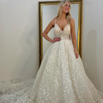 A woman modeling a white A-line wedding dress with a sweetheart neckline, thin spaghetti straps, and a full lace-embroidered skirt with a train, standing in front of a gold-framed mirror.