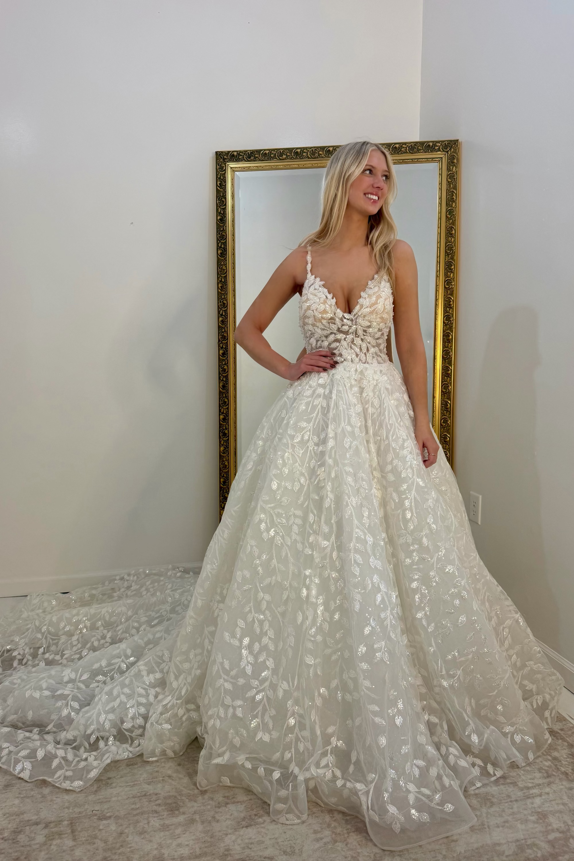 A woman modeling a white A-line wedding dress with a sweetheart neckline, thin spaghetti straps, and a full lace-embroidered skirt with a train, standing in front of a gold-framed mirror.