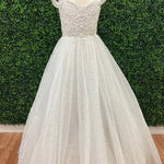 White tulle ball gown dress with a lace bodice and cap sleeves, displayed on a mannequin in front of a green hedge wall and wood floor.
