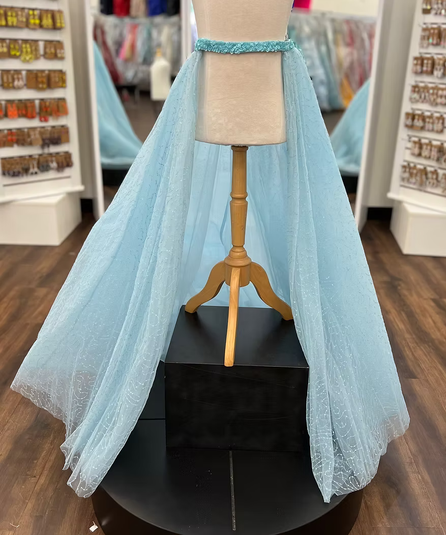 A light blue glitter tulle overskirt is displayed on a cream mannequin with a wooden stand, set on a black pedestal in a retail store.