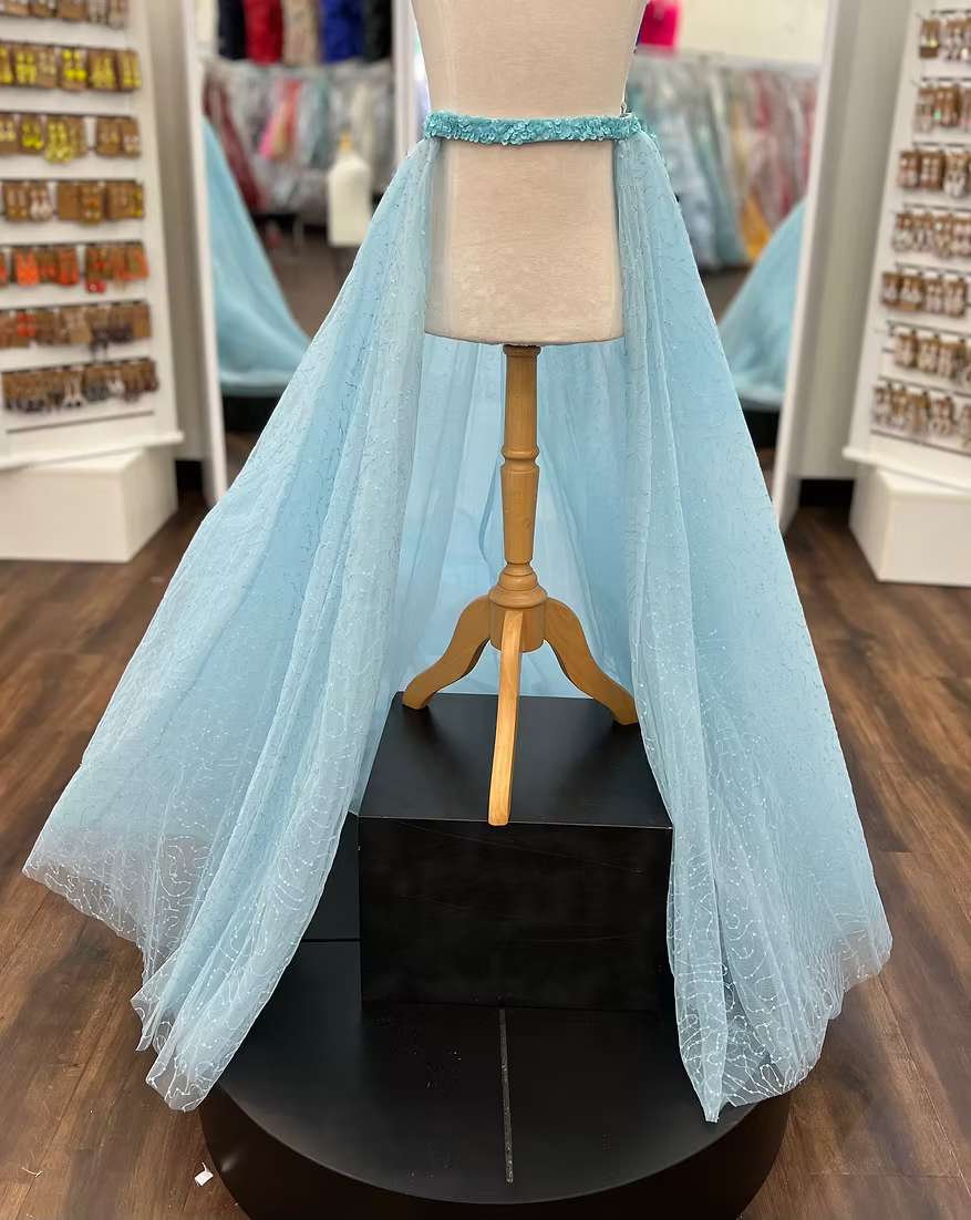 A light blue glitter tulle overskirt is displayed on a cream mannequin with a wooden stand, set on a black pedestal in a retail store.