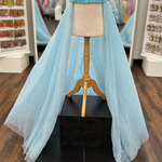 A light blue glitter tulle overskirt is displayed on a cream mannequin with a wooden stand, set on a black pedestal in a retail store.
