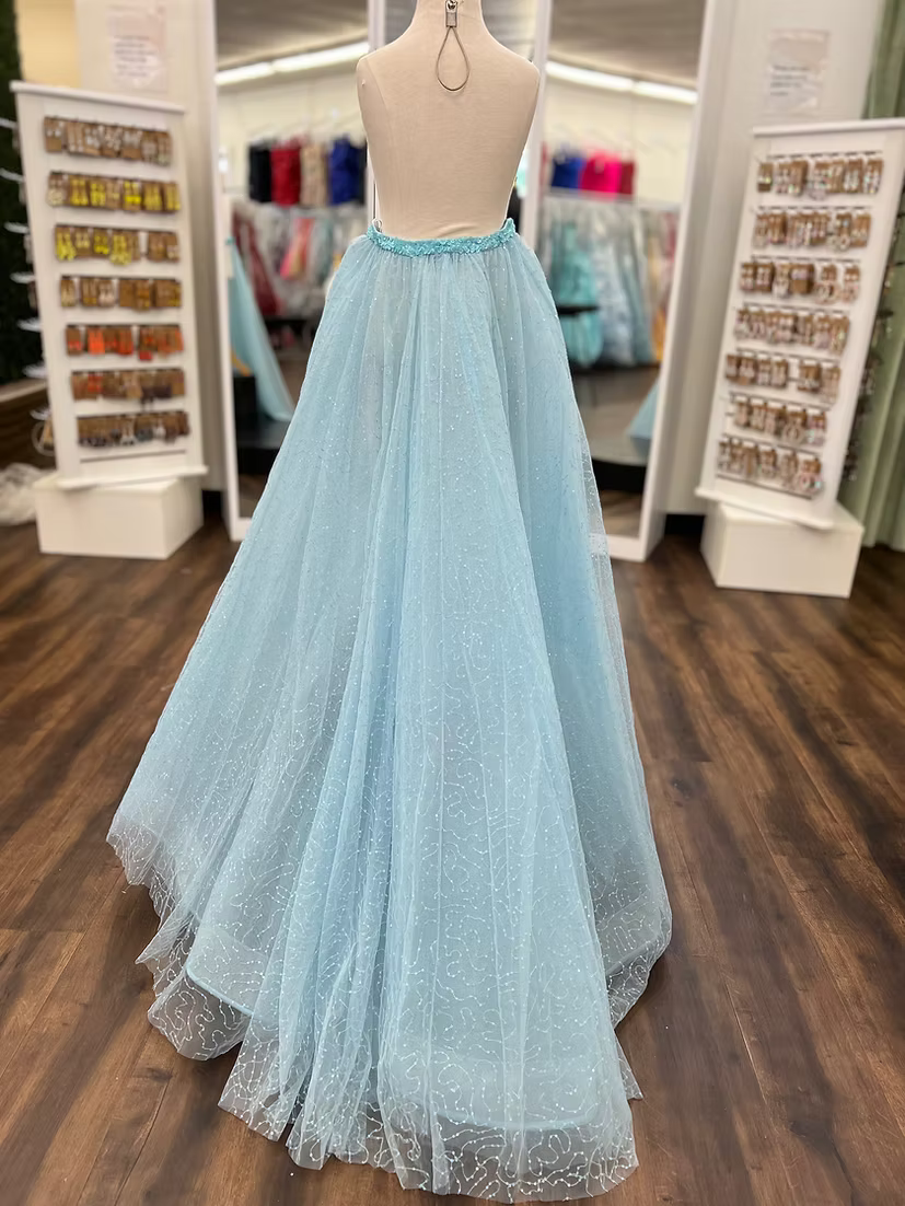 A light blue tulle ball gown displayed on a dress form mannequin inside a clothing boutique. The dress features a fitted bodice with a light blue beaded waistband and a voluminous, sparkly tulle skirt.