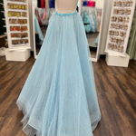 A light blue tulle ball gown displayed on a dress form mannequin inside a clothing boutique. The dress features a fitted bodice with a light blue beaded waistband and a voluminous, sparkly tulle skirt.