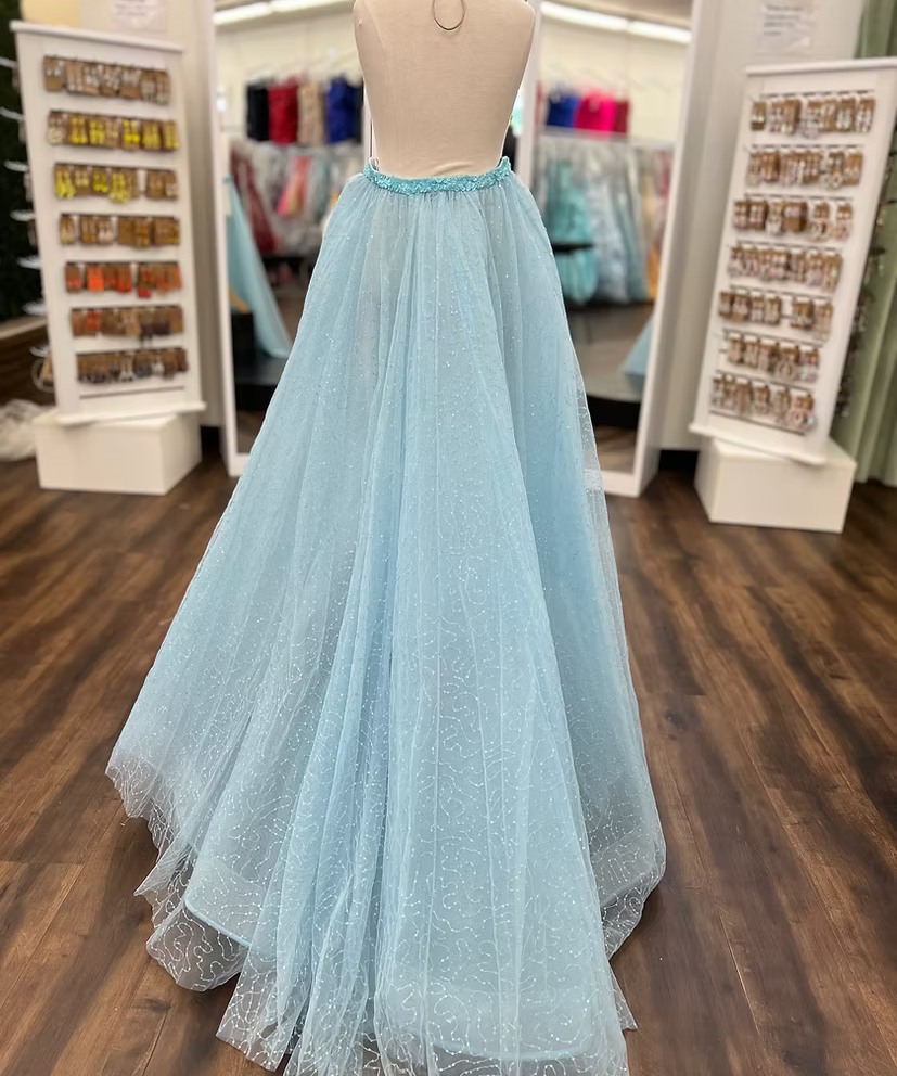 A light blue tulle ball gown displayed on a dress form mannequin inside a clothing boutique. The dress features a fitted bodice with a light blue beaded waistband and a voluminous, sparkly tulle skirt.