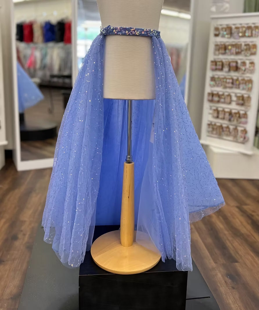 A periwinkle tulle overskirt displayed on a mannequin torso stand in a retail store. The garment has a sparkly blue sequin waistband and a layered tulle skirt with small reflective glitter-like dots.