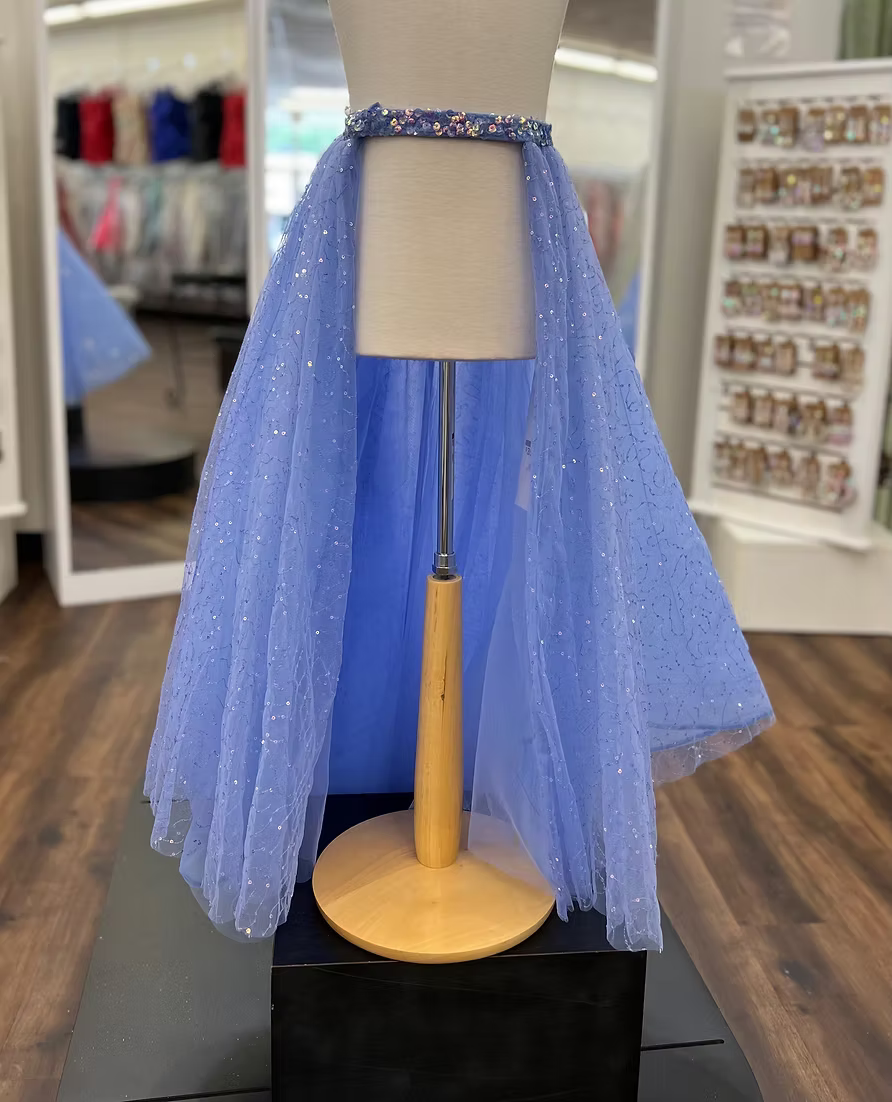 A periwinkle tulle overskirt displayed on a mannequin torso stand in a retail store. The garment has a sparkly blue sequin waistband and a layered tulle skirt with small reflective glitter-like dots.