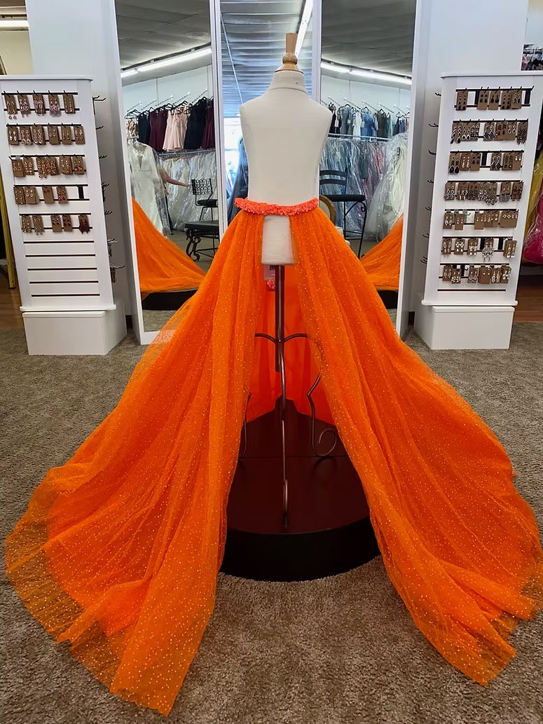 A bright orange tulle overskirt displayed on a mannequin in a formalwear boutique. The overskirt features a dramatic, floor-length train with subtle dotted sparkle details and a sequin covered waistband.
