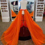 A bright orange tulle overskirt displayed on a mannequin in a formalwear boutique. The overskirt features a dramatic, floor-length train with subtle dotted sparkle details and a sequin covered waistband.