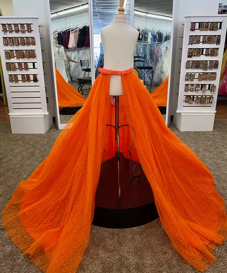A bright orange tulle overskirt displayed on a mannequin in a formalwear boutique. The overskirt features a dramatic, floor-length train with subtle dotted sparkle details and a sequin covered waistband.