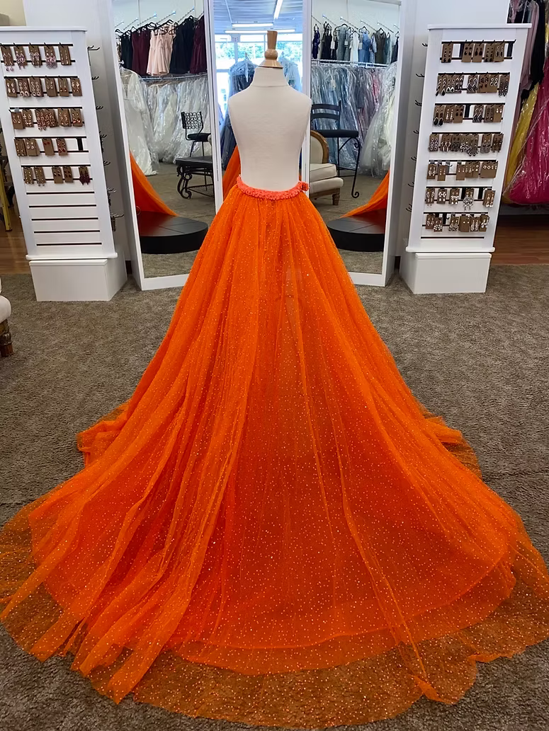 A bright orange tulle overskirt displayed on a mannequin in a formalwear boutique. The overskirt features a dramatic, floor-length train with subtle dotted sparkle details and a sequin covered waistband.
