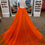 A bright orange tulle overskirt displayed on a mannequin in a formalwear boutique. The overskirt features a dramatic, floor-length train with subtle dotted sparkle details and a sequin covered waistband.
