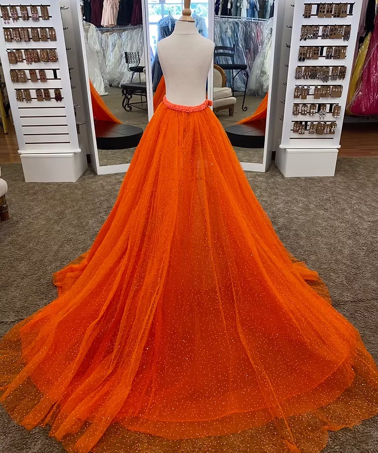 A bright orange tulle overskirt displayed on a mannequin in a formalwear boutique. The overskirt features a dramatic, floor-length train with subtle dotted sparkle details and a sequin covered waistband.