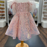 A pink sequined girl's party dress with feathered puff sleeves, displayed on a wooden dress form mannequin inside a retail store.