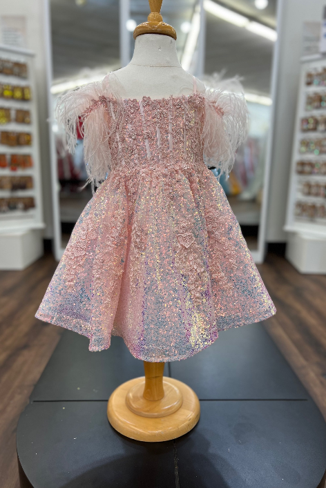 A pink sequined girl's party dress with feathered puff sleeves, displayed on a wooden dress form mannequin inside a retail store.
