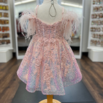 A pink iridescent sequined baby dress with feathered off-the-shoulder sleeves, displayed on a wooden dress form mannequin inside a retail store.