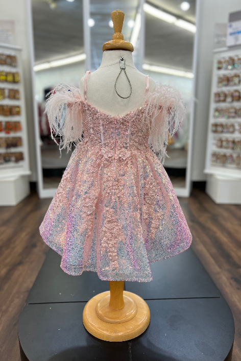 A pink iridescent sequined baby dress with feathered off-the-shoulder sleeves, displayed on a wooden dress form mannequin inside a retail store.