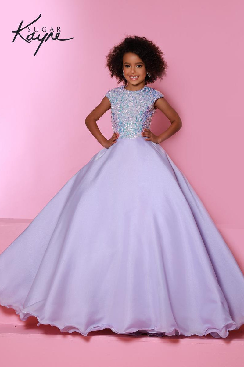 A young girl with curly dark hair models a lavender purple ball gown with a sparkling silver sequined bodice, posing against a solid pink background. The brand logo 'Sugar Kayne' is visible in the top left corner.