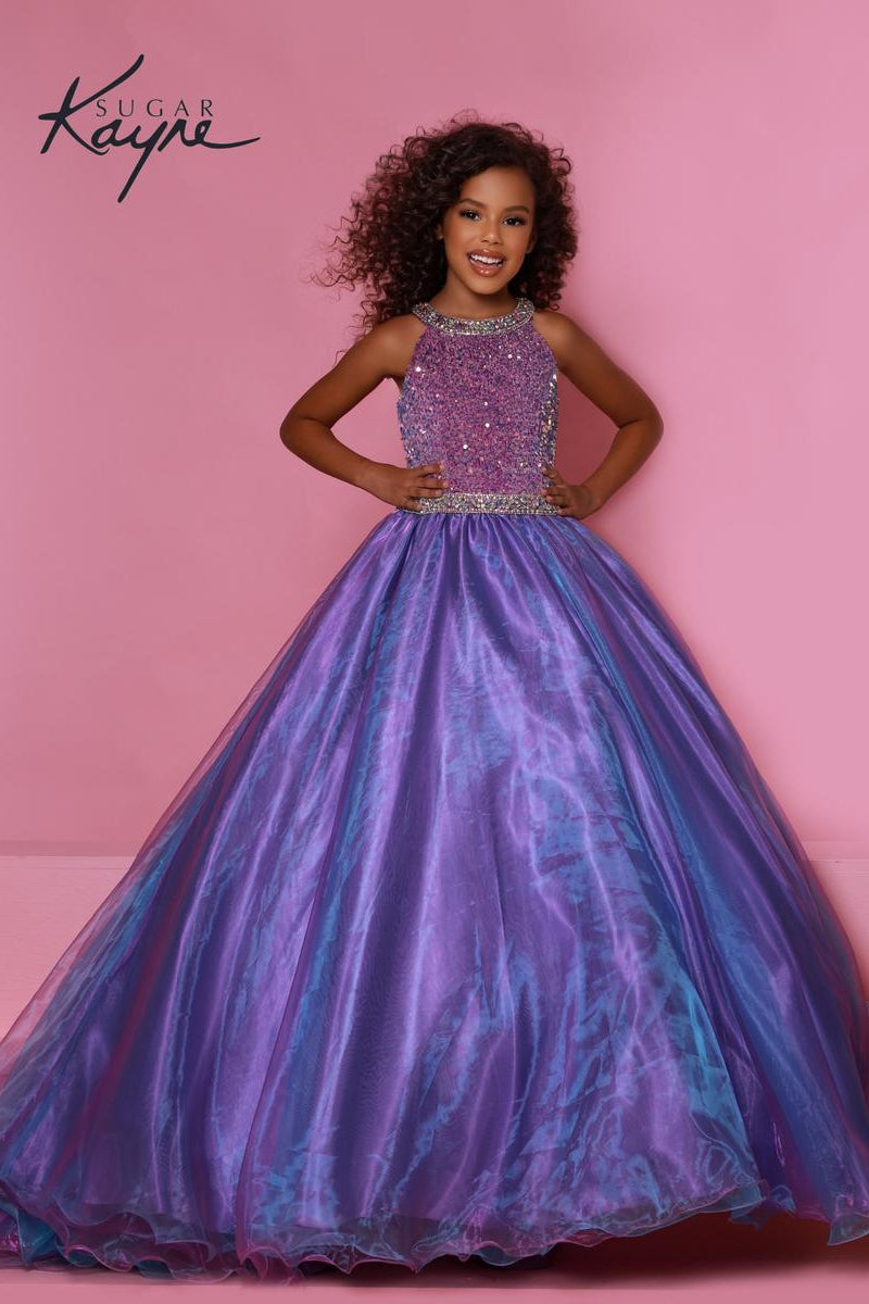 A young girl models a Sugar Kayne ball gown with a purple and blue iridescent tulle skirt and a sparkling beaded bodice, set against a solid pink background.