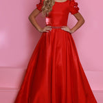 A young woman modeling a bright red formal ball gown with puffed short sleeves, a fitted bodice, and a full satin-like skirt against a pink studio backdrop. The Sugar Kayne logo appears in the upper left corner.