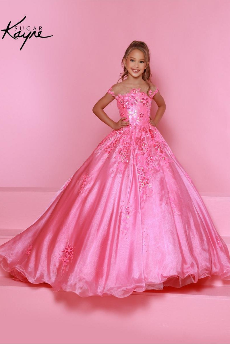 A young girl modeling a bright pink off-the-shoulder ball gown with floral lace applique, standing against a solid pink studio backdrop. The Sugar Kaye brand logo is visible in the top left corner.