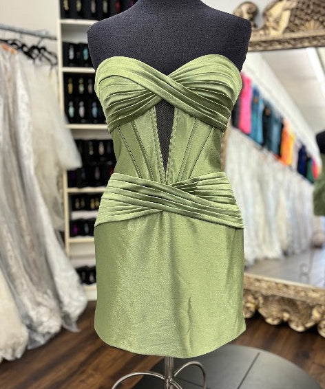 A strapless sage green mini dress with a sweetheart neckline and twisted waist details, displayed on a black dress form mannequin inside a bridal shop.