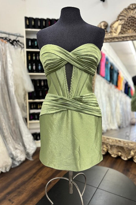 A strapless sage green mini dress with a sweetheart neckline and twisted waist details, displayed on a black dress form mannequin inside a bridal shop.
